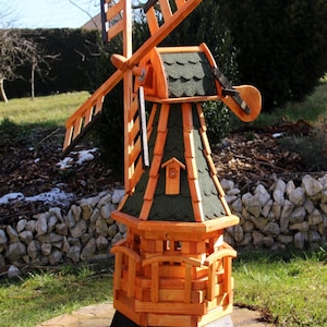 Puede incluir: Un molino de viento de madera con detalles en naranja y negro. Las aspas están anguladas y el tejado está cubierto de tejas verdes. El molino está sobre una base de piedra en una zona de césped, con un muro de piedra al fondo.