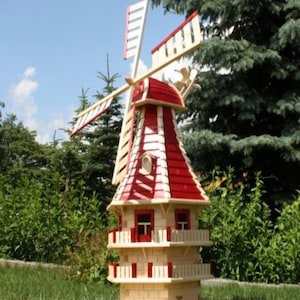 Op de afbeelding: Een houten vogelhuisje in de vorm van een windmolen, met een rood dak en witte accenten. Het vogelhuisje heeft meerdere niveaus met kleine balkons en ramen. De wieken van de molen zijn wit en rood. Het staat in een tuin.