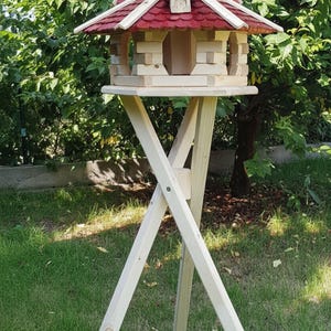 May include: A wooden birdhouse with a red shingled roof and a light-colored wooden body. The birdhouse is mounted on a three-legged stand. The birdhouse has a small, arched doorway and is surrounded by green grass and trees.
