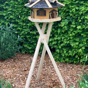 May include: A wooden birdhouse with a dark shingled roof and multiple window openings. The birdhouse is mounted on a tall, three-legged wooden stand. The birdhouse is surrounded by green foliage and mulch.