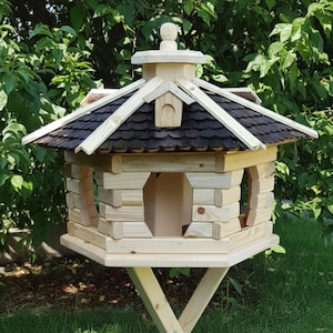 May include: A wooden birdhouse with a multi-sided design, featuring a dark brown shingled roof and light-colored wooden walls. The birdhouse has arched openings and is supported by a wooden stand. The background includes green foliage.