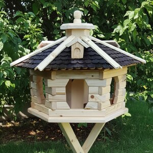 May include: A wooden birdhouse with a multi-sided design, featuring a dark brown shingled roof and light-colored wood construction. The birdhouse has arched openings and is supported by a wooden stand. The background includes green foliage.
