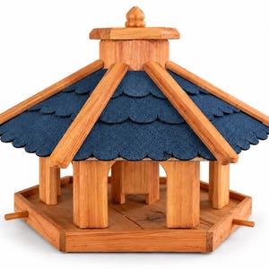 Op de afbeelding: Een zeshoekige houten vogelvoederbak met een natuurlijke lichtbruine houtafwerking, een centrale steunstructuur en een donkerblauw geschulpt dak met een kleine decoratieve knop op de top. De basis bevat kleine houten zitstokken aan de zijkanten.