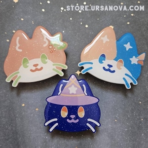 May include: Three enamel pins featuring cartoon cats with different designs. The first pin is a light orange cat with green eyes and a small red berry on its head. The second pin is a white cat with blue and orange ears and a star on its head. The third pin is a dark blue cat wearing a purple hat with a star on top. All three pins have glitter embedded in the enamel.