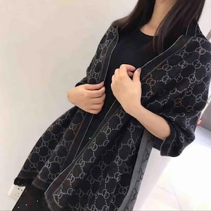 May include: A dark gray scarf with a repeating pattern of interlocking letters. The scarf has a contrasting border and is draped over the shoulders. The scarf is made of a soft material.