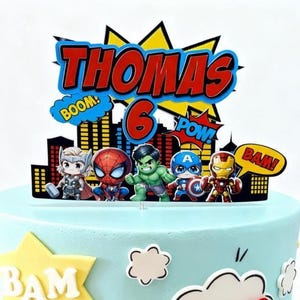 Puede incluir: Una tarta azul claro con temática de superhéroes. La decoración de la tarta muestra el nombre "Thomas" y el número "6", junto con exclamaciones de cómic y figuras de superhéroes como Thor y Spider-Man.
