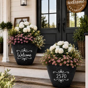 May include: Two black textured flower pots with white script. One pot says "Welcome" and the other says "2570". Each pot is filled with white and pink flowers. The pots are on a wooden porch.