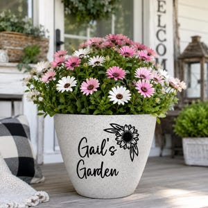 Op de afbeelding: Een grote, gespikkelde grijze bloempot gevuld met roze en witte madeliefachtige bloemen. De pot heeft de tekst "Gail's Garden" en een zonnebloemontwerp. De pot staat op een houten oppervlak.