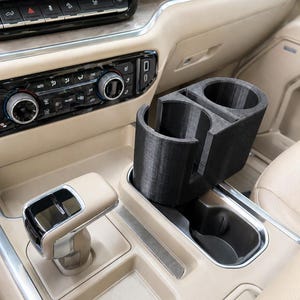 May include: Black 3D-printed car cup holder with two circular openings and a rectangular section. The cup holder is designed to fit into a car's center console, next to the gear shift.
