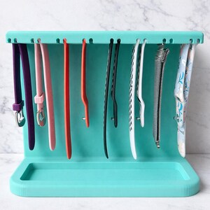 May include: A turquoise watch band organizer with various colored watch bands hanging from it. The bands are purple, pink, red, black, white, and blue. The organizer has a tray at the bottom.