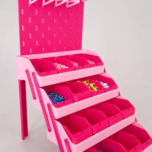 Op de afbeelding: Een gelaagde roze opbergorganizer met een verticaal pegboard-achterpaneel met lichtroze haken, vier rijen open bakken in verschillende tinten roze en kleine kleurrijke voorwerpen die in de vakken zijn opgeborgen voor organisatie.