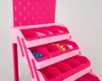 May include: A tiered pink storage organizer featuring a vertical pegboard back panel with light pink hooks, four rows of open-front bins in varying shades of pink, and small colorful items stored within the compartments for organization.