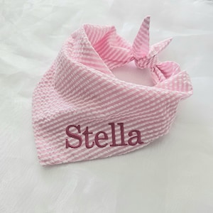 Peut inclure: Un bandana triangulaire pour animal de compagnie en tissu seersucker rayé rose et blanc. Le prénom Stella est brodé en fil rose foncé sur le devant. Le tissu est noué au coin supérieur pour permettre une fixation sécurisée autour du cou.