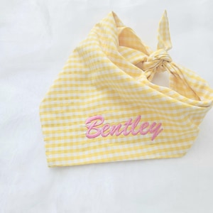 May include: A yellow and white gingham patterned pet bandana featuring the name Bentley embroidered in pink script lettering. The fabric is tied in a knot at the top corner, creating a triangular shape with a soft, folded texture throughout.
