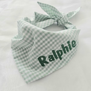 May include: A light green and white gingham dog bandana with the name "Ralphie" embroidered in dark green. The bandana is tied at the top in a bow.