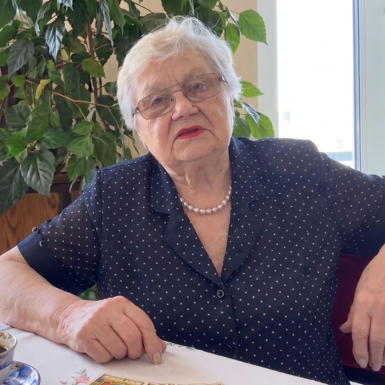 Puede incluir: Una persona mayor de pelo blanco corto viste una blusa azul marino de manga corta con estampado de lunares blancos, un collar de perlas de una sola hilera y gafas de montura rectangular con bordes met&aacute;licos finos, frente a una planta.
