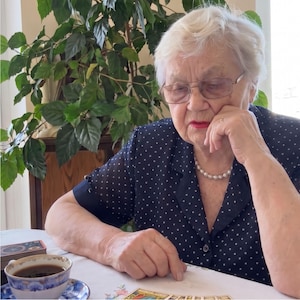 Puede incluir: Una persona mayor con cabello blanco corto y gafas viste una blusa azul marino de manga corta con un patr&oacute;n de lunares blancos y un collar de perlas. Est&aacute; sentada a una mesa con una taza de caf&eacute; negro y una baraja de cartas del tarot.