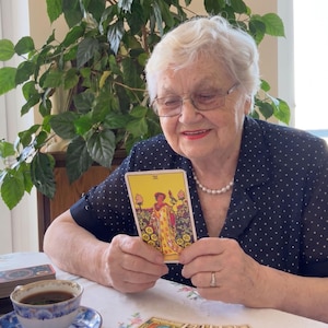 Puede incluir: Una carta de tarot rectangular con fondo amarillo y una ilustraci&oacute;n de una figura de pie en un jard&iacute;n con nueve monedas de oro. La carta es sostenida por manos, con una taza de l&iacute;quido oscuro y una baraja de cartas visibles sobre la mesa.