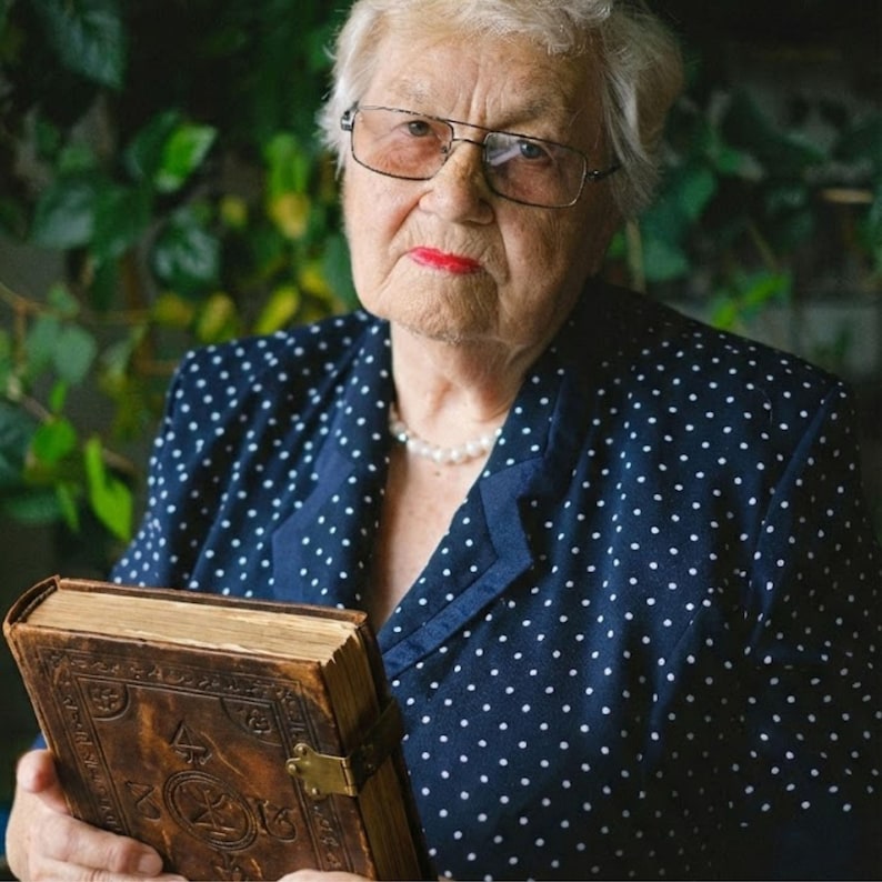 May include: An elderly person wearing glasses and a navy blue blazer with white polka dots holds a vintage, leather-bound book. The book has a brass clasp and ornate detailing. A pearl necklace is also visible.
