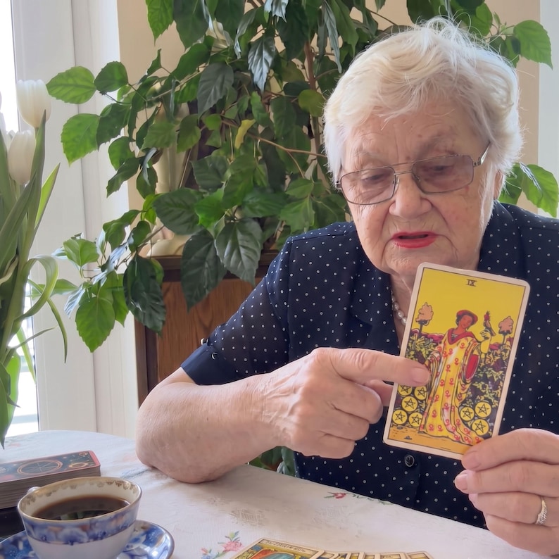 Puede incluir: Una carta de tarot rectangular con la ilustraci&oacute;n del Nueve de Oros, que muestra a una figura con una t&uacute;nica amarilla de pie en un jard&iacute;n con nueve monedas de oro. La carta se sostiene en alto, mostrando el n&uacute;mero romano IX en la parte superior central.