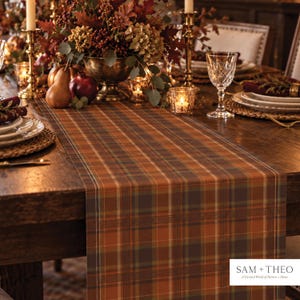 May include: A rectangular table runner featuring a plaid pattern in shades of orange, brown, and dark green. The fabric displays a grid of intersecting lines. A white logo in the bottom right corner reads SAM + THEO A Curated World of Pattern + Home.