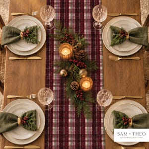 May include: A dining table setting with a burgundy and white plaid runner, four white plates with olive green napkins, gold napkin rings, red berries, pine needles, pinecones, two lit candles, and the text SAM + THEO A Curated World of Pattern + Home.