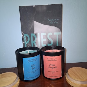 May include: Two black candle jars with labels reading "Tyler Bell" and "Poppy Danforth" sit in front of a book titled "PRIEST". The candles have wooden lids. The Tyler Bell candle is blue, and the Poppy Danforth candle is pink.