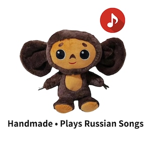 May include: A handmade plush toy in the shape of a cartoon character with large ears and a smiling face. The toy is brown and tan with large black eyes. The text "Handmade • Plays Russian Songs" is below the toy.