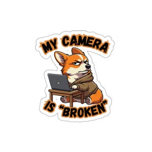 Puede incluir: Una pegatina con un corgi de dibujos animados con una sudadera con capucha marrón, sentado en una pequeña mesa de madera con un portátil. El texto "MY CAMERA IS BROKEN" está en naranja y negro.
