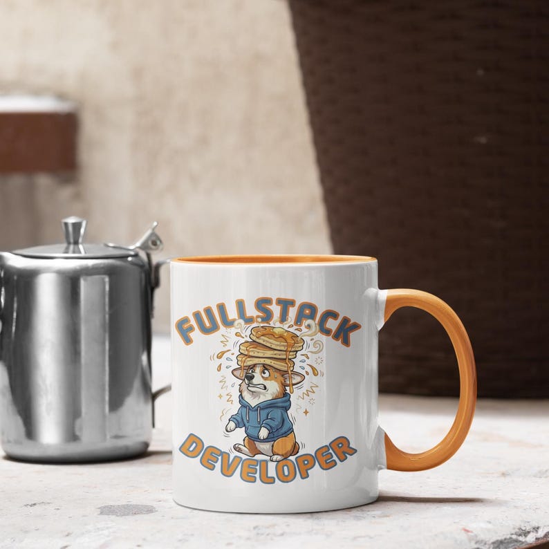 May include: White ceramic mug with an orange handle and interior. The mug features a cartoon corgi wearing a blue hoodie and a stack of pancakes on its head. The words "FULLSTACK DEVELOPER" are printed on the mug.