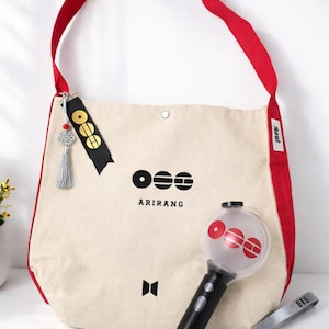 Pode incluir: Bolsa tote creme com alças vermelhas, com logotipo preto e texto "ARIRANG". Inclui um bastão de luz preto e vermelho e uma pulseira cinza com texto "BTS".