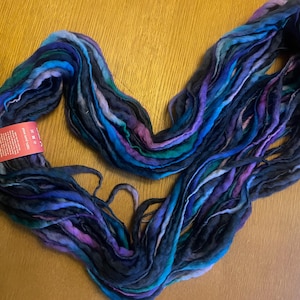 May include: A skein of thick, unspun roving wool yarn featuring a variegated colour palette of deep navy blue, royal blue, teal, and vibrant purple. A rectangular red product tag with white text is attached to the side of the fibre bundle.