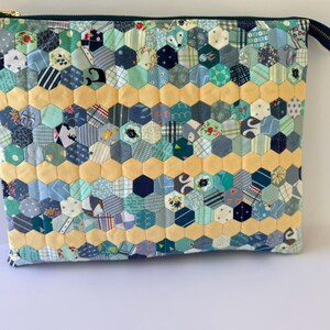 Könnte beinhalten: Gesteppte Tasche mit Patchwork-Design. Die Tasche hat ein Wabenmuster aus verschiedenen blauen, grünen und gemusterten Stoffen. Ein Band aus gelben Sechsecken verläuft über die Mitte. Sie hat einen dunkelgrünen Reißverschluss und einen goldfarbenen Reißverschluss.