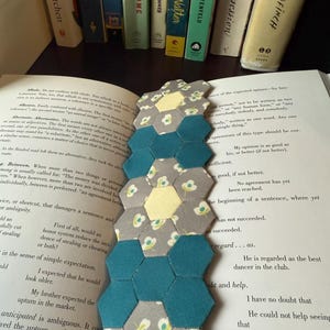 May include: A handmade fabric bookmark with a honeycomb design. The bookmark features alternating teal, gray floral, and yellow hexagonal fabric pieces, placed on an open book with a row of books in the background.
