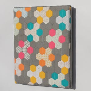 May include: A gray fabric item with a quilted hexagonal pattern in various colors. The hexagons are in shades of yellow, orange, pink, teal, and white. The item has a gray border and appears to be a book cover or a similar textile product.