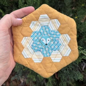 May include: A handmade, quilted, hexagonal trivet with a fox design. The trivet features a mustard yellow border and a central design of blue and white patterned hexagons surrounding a fox face. The trivet is approximately 6 inches wide.