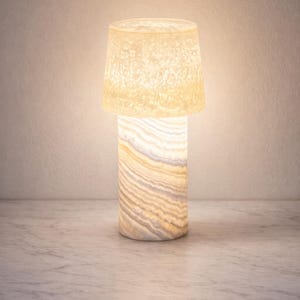 Può includere: Lampada da tavolo cilindrica con paralume strutturato bianco sporco e base in pietra traslucida a strati. La lampada emette una calda luce, ideale per l'illuminazione d'ambiente. La base è alta circa 20 cm.
