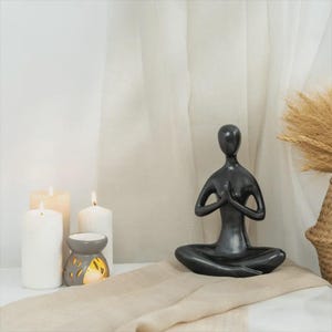 May include: A dark grey yoga figurine in a seated meditation pose, with hands clasped in prayer. The figurine is on a beige cloth, with lit candles and an essential oil diffuser in the background. A woven basket with dried plants is on the right.