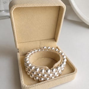 May include: A multi-strand bracelet composed of lustrous white spherical beads, arranged in three circular rows. The bracelet is displayed inside an open, square-shaped beige velvet jewellery box with a soft, light-coloured interior lining.