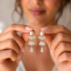 Puede incluir: Un par de pendientes colgantes en tono dorado con un perno de perla blanca en la parte superior, seguido de tres secciones escalonadas en forma de abanico compuestas por cristales transparentes de corte baguette que disminuyen de tamaño.