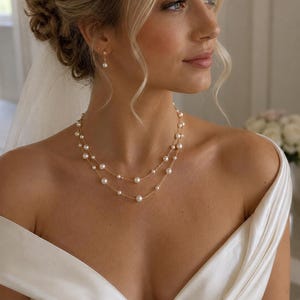 May include: A double-strand gold chain necklace featuring evenly spaced white pearls, paired with matching single pearl drop earrings. The jewelry set is worn with an off-the-shoulder white satin garment and a sheer veil, highlighting the delicate metallic links and round pearl accents.