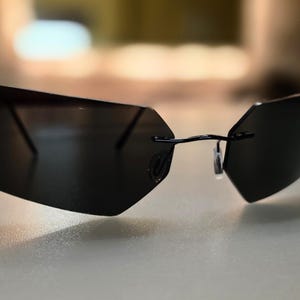 Puede incluir: Gafas de sol negras sin montura con un diseño geométrico. Las lentes son oscuras y la montura es fina y negra. Las gafas de sol tienen una estética moderna y minimalista.
