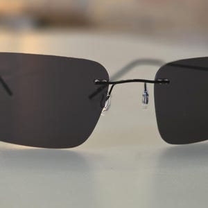 Puede incluir: Gafas de sol sin montura con lentes de color gris oscuro y una fina montura negra. Las lentes rectangulares están conectadas por un fino puente y patillas. Las gafas de sol están diseñadas para un aspecto elegante y moderno.