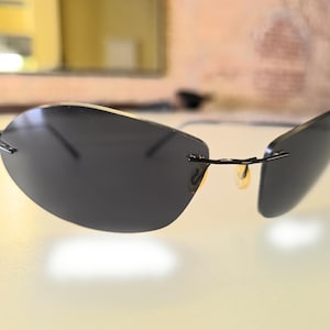 Puede incluir: Gafas de sol sin montura con lentes de color gris oscuro y finas monturas de metal negro. Las gafas de sol tienen un diseño elegante y moderno con un pequeño puente nasal dorado. Las lentes son de forma ovalada.