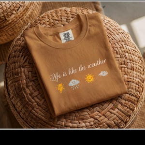 May include: A tan Comfort Colors t-shirt with the phrase "Life is like the weather" and weather-related graphics. The design includes a lightning bolt, rain cloud, sun, and a cloud. The shirt is folded on a woven wicker surface.