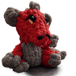 Puede incluir: Un peluche de dragón hecho a mano con ganchillo en rojo y gris. El dragón tiene un aspecto texturizado y grueso con ojos negros y una nariz negra. El peluche está sentado, con los brazos y las piernas visibles.