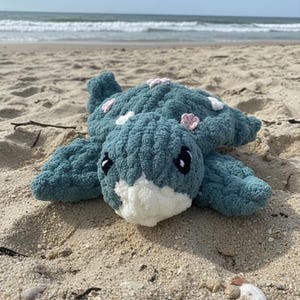 Könnte beinhalten: Ein gehäkeltes Schildkröten-Plüschtier in Türkis mit weißen und rosa Akzenten, das auf einem Sandstrand liegt. Die Schildkröte hat schwarze Augen und ein weißes Gesicht. Das Meer und der Himmel sind im Hintergrund zu sehen.