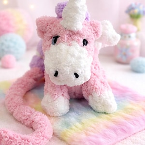 May include: A plush pink unicorn toy with a white horn, mane, and hooves. The unicorn sits on a rainbow-colored blanket. The toy has black eyes and a long, curved tail. The background is soft pink.
