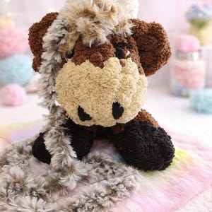 May include: A plush stuffed animal shaped like a cow with brown and dark brown textured yarn, a cream-coloured muzzle, and black nostrils. It features a long, shaggy, multi-toned faux fur scarf draped over its head and body, with white tufts.