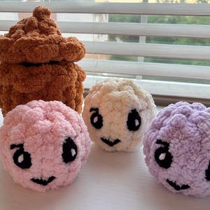 May include: A set of four crocheted plush items including one brown acorn-shaped piece and three round, textured spheres in light pink, cream, and light purple. Each sphere features embroidered black eyes and a small black mouth on the front.
